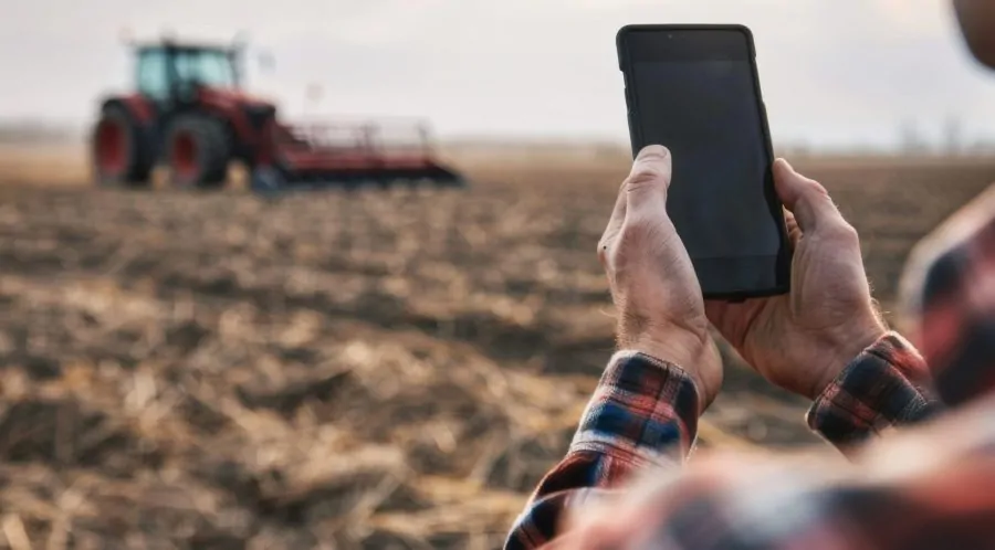 Modern farmer holding phone on the field, blurred tractor in the background